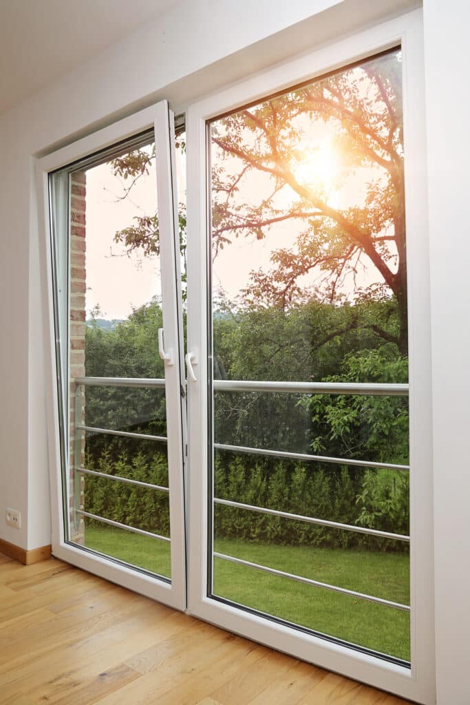 Intérieur lumineux avec sol en parquet clair ouvrant sur un jardin verdoyant via une porte-fenêtre oscillo-battante PVC blanche.