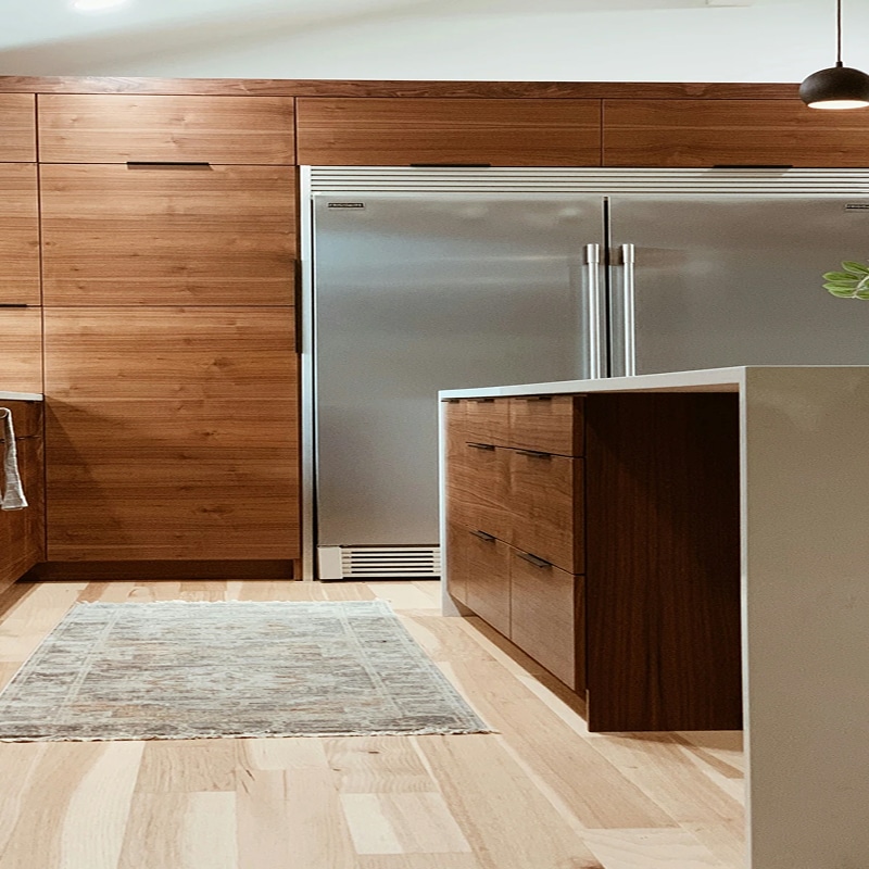 Cuisine moderne avec armoires en bois de noyer, réfrigérateur Sub-Zero en acier inoxydable et îlot blanc avec tiroirs assortis.